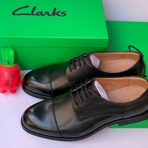 A pair of black Clark's Loafer shoes, displayed on a green box with a small red plush toy.